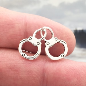 May include: Silver-toned metal charm in the shape of handcuffs. The charm is small and detailed, with a realistic design.