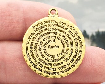 May include: Gold-toned metal charm with a circular design featuring the Lord's Prayer in Spanish. The charm is engraved with the text "Padre nuestro, que estás en el cielo, santificado sea tu nombre; venga a nosotros tu reino; hágase tu voluntad, como en el cielo, así también en la tierra. Danos hoy nuestro pan de cada día; y perdónanos nuestras deudas, como también nosotros perdonamos a nuestros deudores. Y no nos dejes caer en la tentación, mas líbranos del mal. Amén."