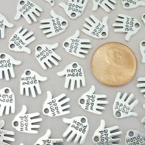May include: Silver-coloured hand-shaped charms with the words "Hand Made" engraved on them. A U.S. penny is included for size comparison. The charms are small, with a hole for stringing or attaching. The charms are scattered across a light grey surface.