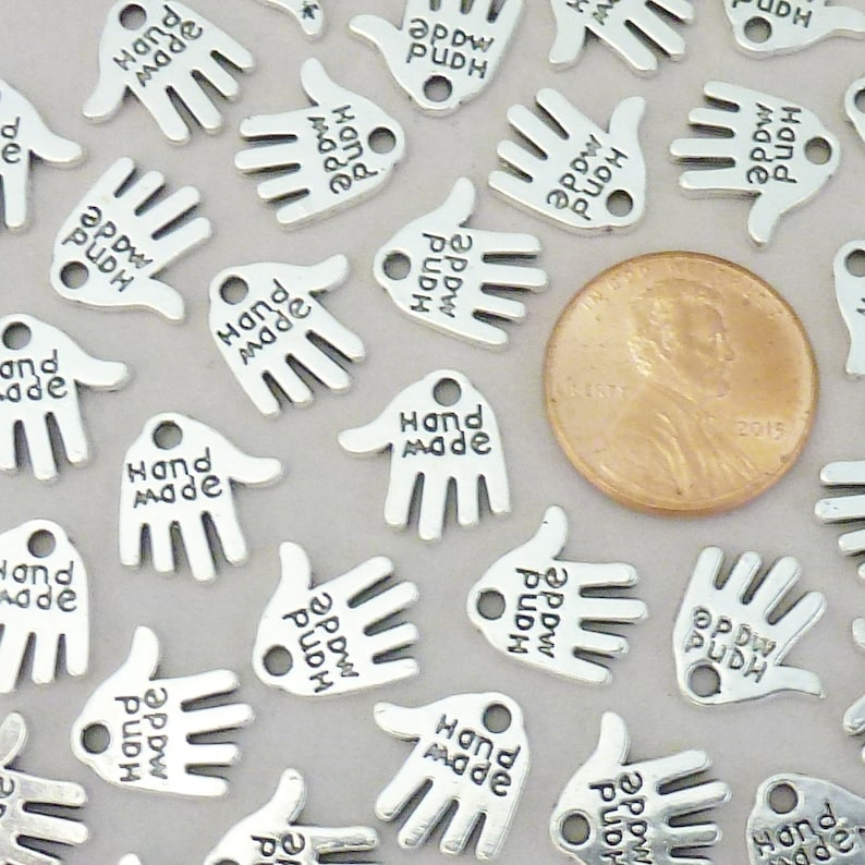 May include: Silver-coloured hand-shaped charms with the words "Hand Made" engraved on them. A U.S. penny is included for size comparison. The charms are small, with a hole for stringing or attaching. The charms are scattered across a light grey surface.
