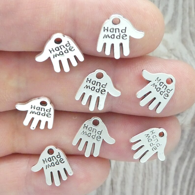 May include: A selection of small, silver-toned hand-shaped charms. Each charm is inscribed with the words "Hand Made". The charms feature a small hole at the top, suitable for attaching to jewellery or craft projects.