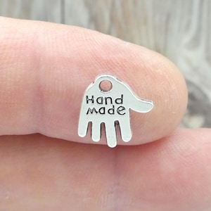 May include: A small, silver-coloured charm in the shape of a hand. The charm has the words "Hand Made" printed on it. The charm has a small hole at the top for stringing.