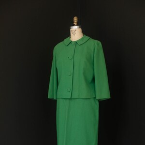 Puede incluir: Un traje de falda verde vintage exhibido en un maniquí. El traje incluye una chaqueta con cuello y tres botones con mangas tres cuartos y una falda a juego hasta la rodilla. El maniquí está sobre un soporte metálico, sobre un fondo negro.