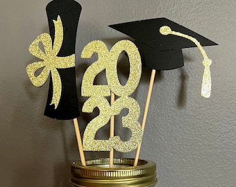 May include: A glass jar filled with black and gold ribbon with a graduation cap, diploma, and gold glitter numbers 2023 on wooden sticks.