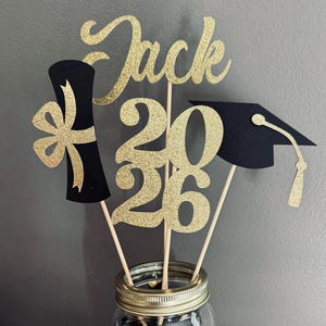 Könnte beinhalten: Ein Abschluss-Tischaufsatz in einem Einmachglas. Er enthält goldglitzernde Ausschnitte mit der Aufschrift "Jack 2026" und Abschlussformen. Das Glas ist mit schwarzem und goldenem Dekofüllmaterial gefüllt und steht auf einer Holzoberfläche.