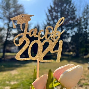 May include: Gold glitter graduation cap topper with the name Jacob and the year 2024. The topper is on a wooden stick and is in front of a vase with pink tulips.