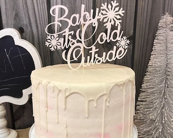 May include: White cake with white frosting and a pink ombre effect. The cake is decorated with a white wooden cake topper that says "Baby It's Cold Outside" with snowflakes.