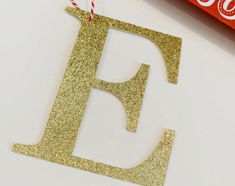 May include: Gold glitter letter E ornament with a red and white string attached.