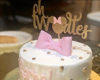 Può includere: Una torta bianca con glassa rosa e spolverata di granella d'oro. La torta è decorata con un fiocco rosa e un topper di glitter dorato che dice "Oh Twodles". La torta ha anche il nome "Rachel" scritto in glassa rosa.