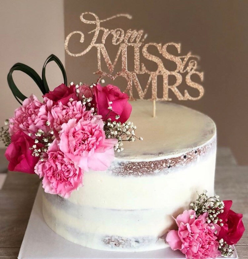 May include: A white cake with pink flowers and a gold glitter topper that says "From Miss to Mrs."