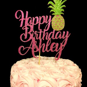 May include: Pink glitter cake topper with the words "Happy Birthday Ashley" and a green glitter pineapple.
