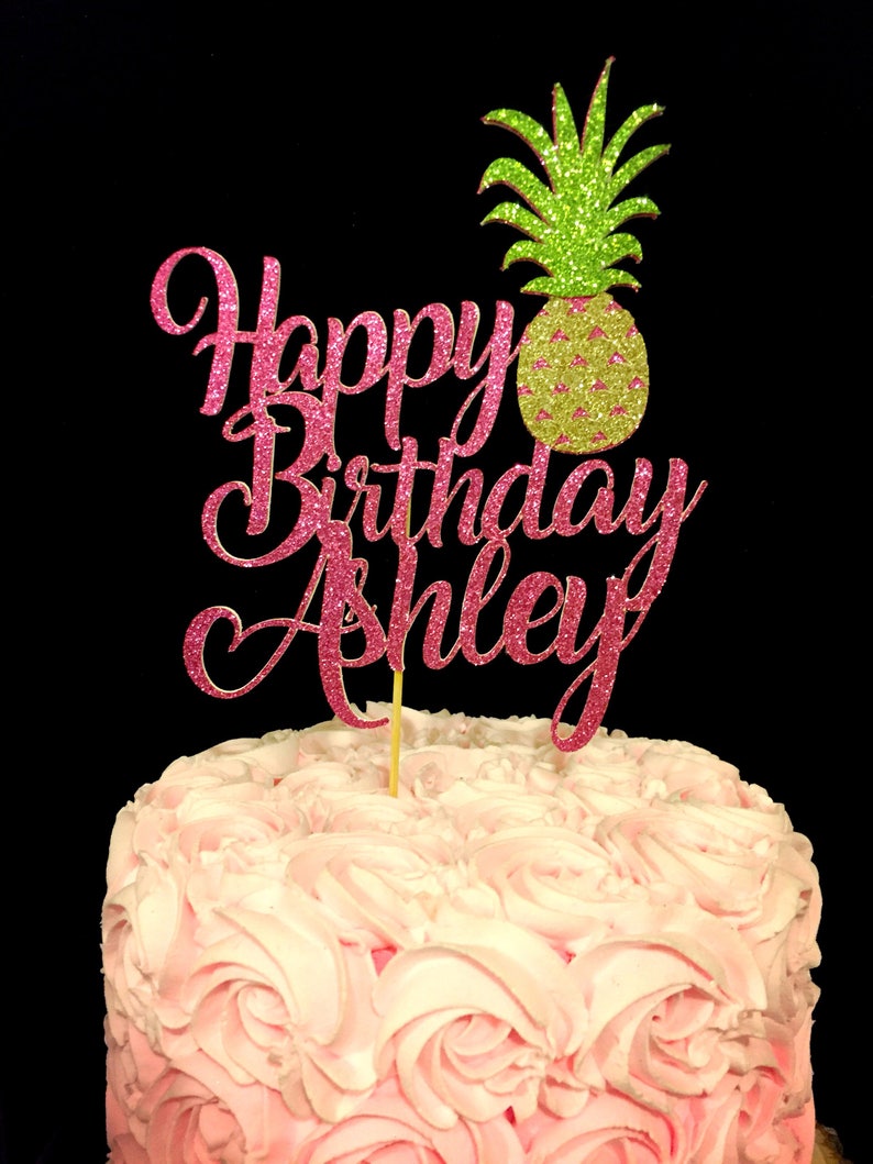May include: Pink glitter cake topper with the words "Happy Birthday Ashley" and a green glitter pineapple.
