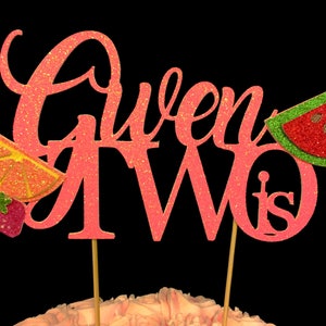 May include: Pink glitter cake topper with the words "Gwen is Two" and a watermelon and orange slice design.