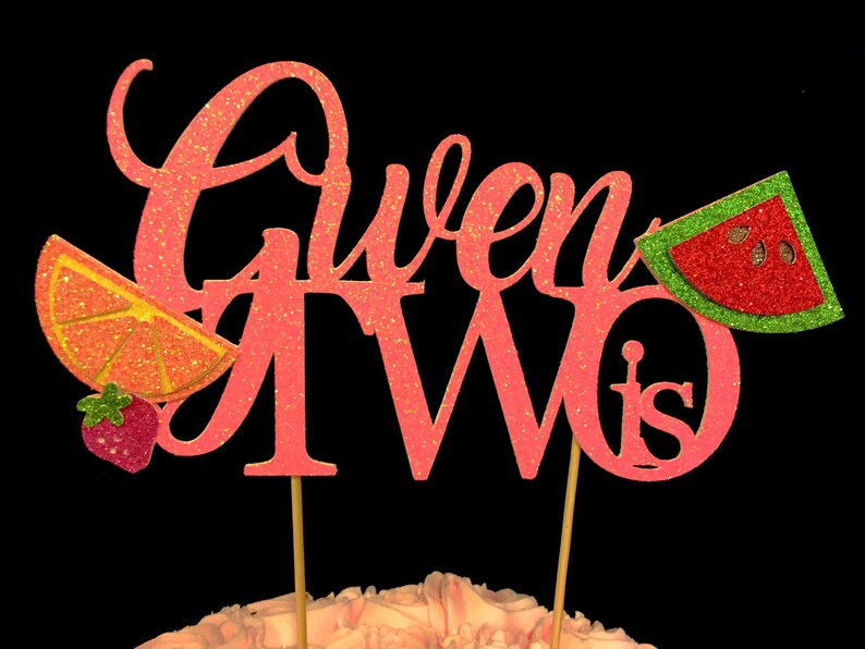 May include: Pink glitter cake topper with the words "Gwen is Two" and a watermelon and orange slice design.