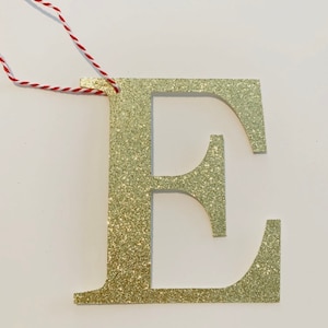 May include: Gold glitter letter E ornament with a red and white string for hanging.