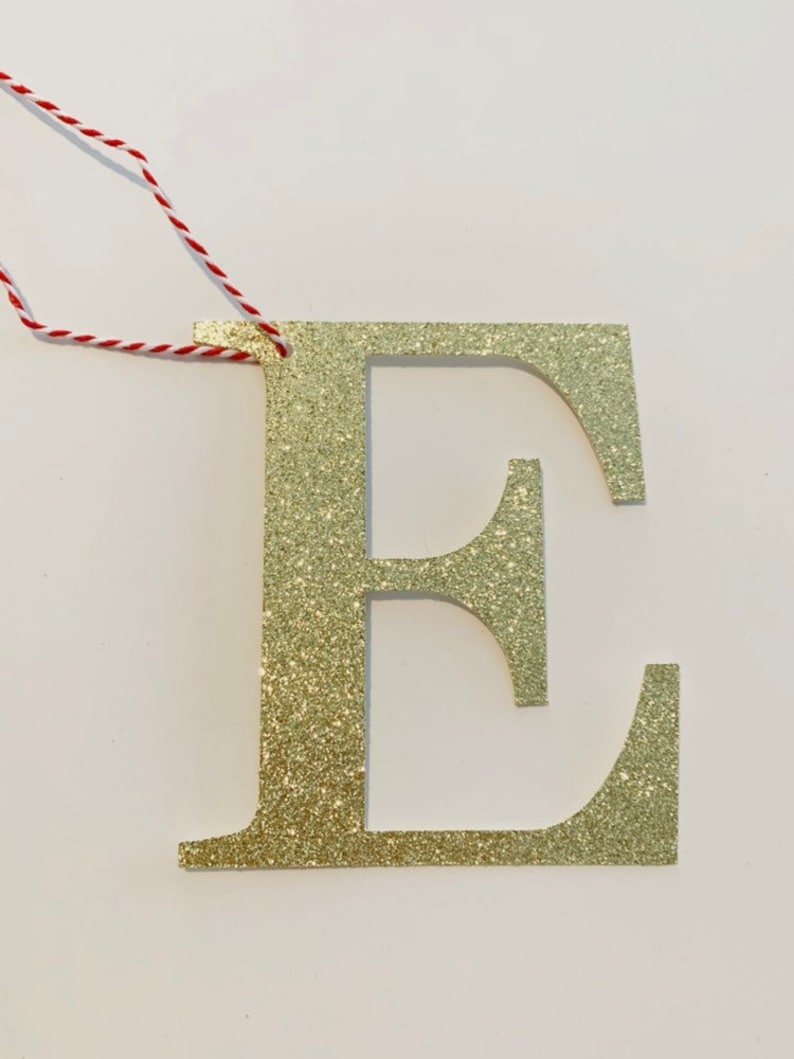 May include: Gold glitter letter E ornament with a red and white string for hanging.