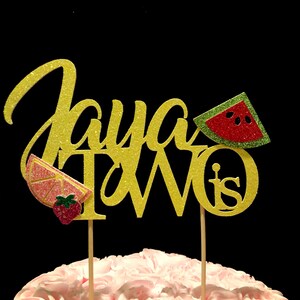 May include: Gold glitter cake topper with the words "Jaya Turns Two" and a watermelon, strawberry, and lemon slice design.