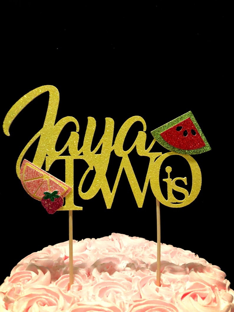 May include: Gold glitter cake topper with the words "Jaya Turns Two" and a watermelon, strawberry, and lemon slice design.