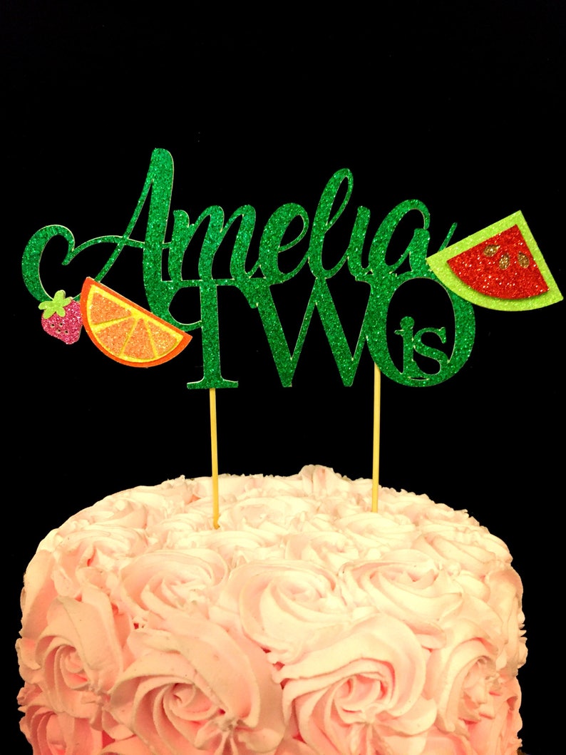May include: Green glitter cake topper with the name Amelia and the number two, with a strawberry, orange slice, and watermelon slice decorations.