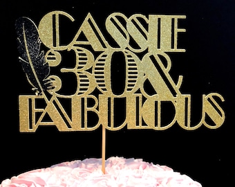 May include: Gold glitter cake topper with the text "Cassie 30 & Fabulous" in a decorative font. A black feather is on the left side of the topper.