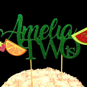 May include: Green glitter cake topper with the name Amelia and the number two, with the word "is" after the number. The topper has a watermelon, orange slice, and strawberry decoration.