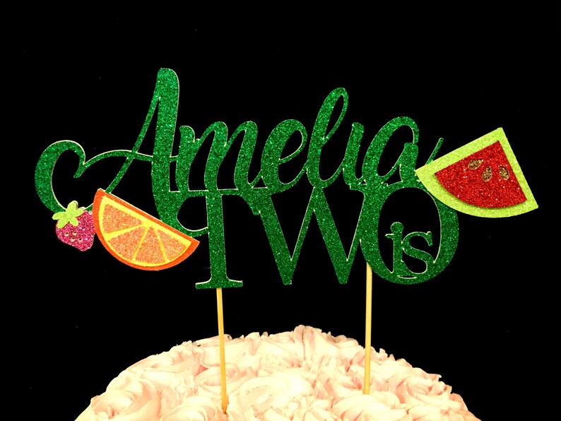 May include: Green glitter cake topper with the name Amelia and the number two, with the word "is" after the number. The topper has a watermelon, orange slice, and strawberry decoration.
