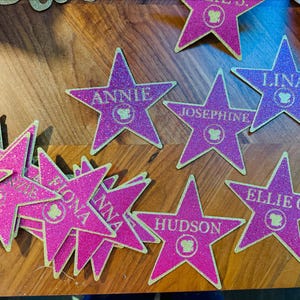 Puede incluir: Decoraciones en forma de estrella rosa brillante con bordes dorados. Cada estrella presenta un nombre, como "Annie" o "Hudson", y un pequeño emblema. Estas estrellas decorativas son adecuadas para fiestas o eventos.