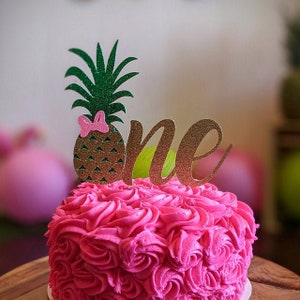 May include: A pink frosted cake with a pineapple and the word "one" on top. The pineapple is green and gold with a pink bow. The cake is decorated with pink frosting in a swirl pattern.