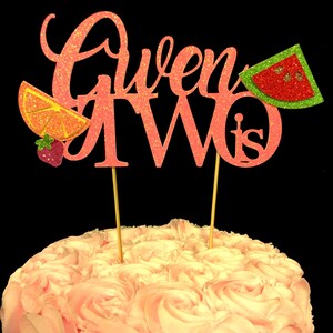 May include: Pink glitter cake topper with the words "Gwen is Two" and a watermelon, orange slice, and strawberry decoration.
