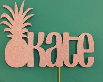 Puede incluir: Topper de pastel de piña de brillo rosa con el nombre "Kate" escrito en brillo rosa.