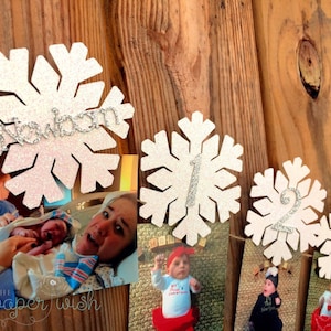 May include: A white snowflake garland with glitter and the word "newborn" on the first snowflake. The garland is decorated with photos of a baby growing up from newborn to one year old.