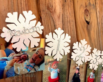 May include: A white snowflake garland with glitter and the word "newborn" on the first snowflake. The garland is decorated with photos of a baby growing up from newborn to one year old.