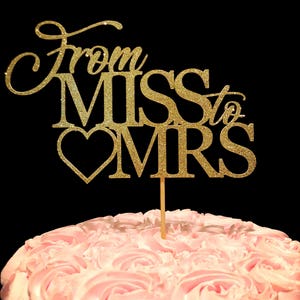 May include: Gold glitter cake topper with the text "From Miss to Mrs" and a heart. The topper is on a pink frosted cake.