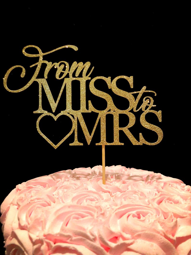 May include: Gold glitter cake topper with the text "From Miss to Mrs" and a heart. The topper is on a pink frosted cake.