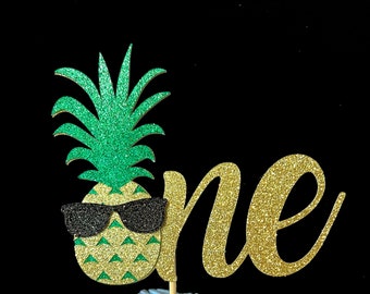 Könnte beinhalten: Goldener Glitzer-Kuchentopper "eins" mit einer grünen Glitzer-Ananas, die eine schwarze Sonnenbrille trägt.