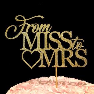 May include: Gold glitter cake topper with the words "From Miss to Mrs" and a heart. The topper is on a pink frosted cake.