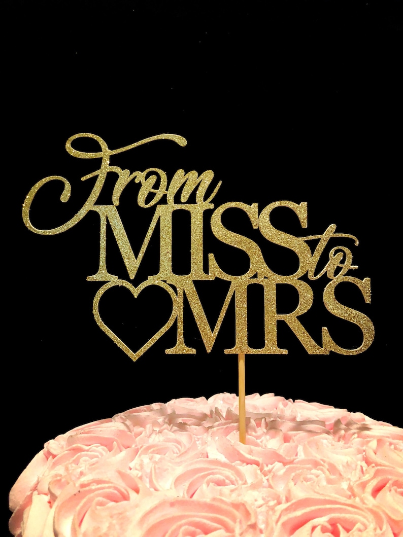 May include: Gold glitter cake topper with the words "From Miss to Mrs" and a heart. The topper is on a pink frosted cake.