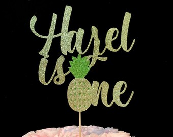 May include: Gold glitter cake topper with the text "Hazel is one" and a green and gold glitter pineapple.