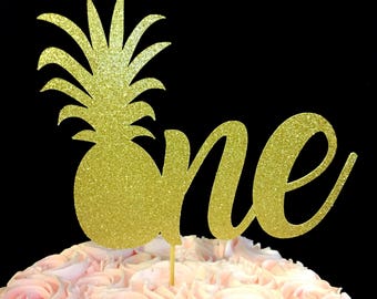 Könnte beinhalten: Goldener Glitzer-Ananas-Kuchentopper mit dem Wort "one" in Kursivschrift. Der Topper befindet sich auf einem rosafarbenen, glasierten Kuchen.