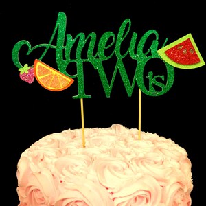 May include: Green glitter cake topper with the name Amelia and the number two, with a strawberry, orange slice, and watermelon slice decorations.