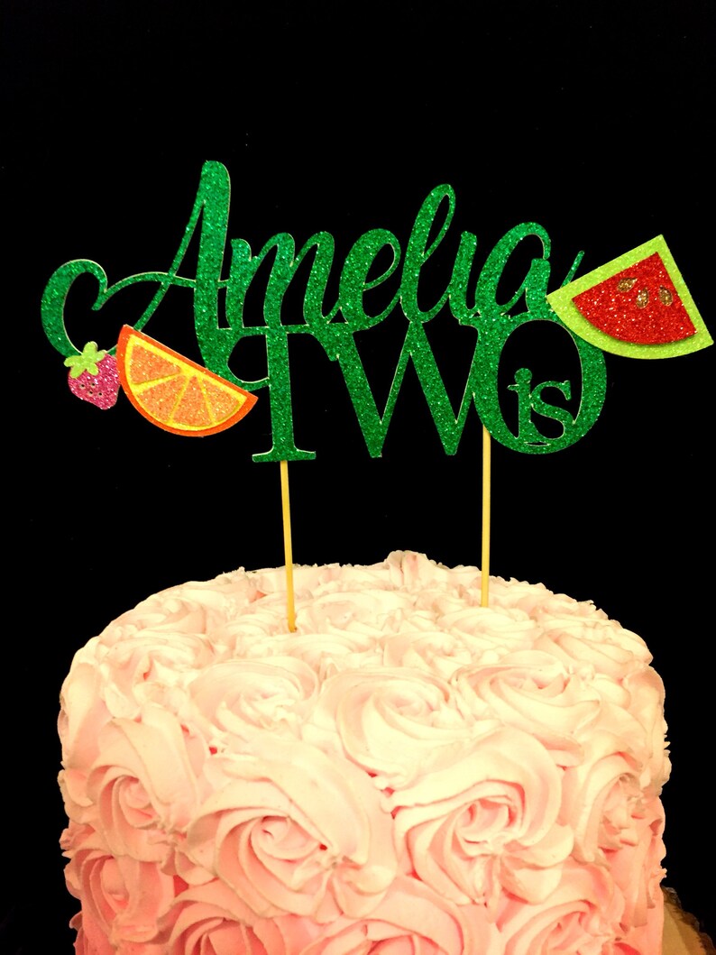 May include: Green glitter cake topper with the name Amelia and the number two, with a strawberry, orange slice, and watermelon slice decorations.