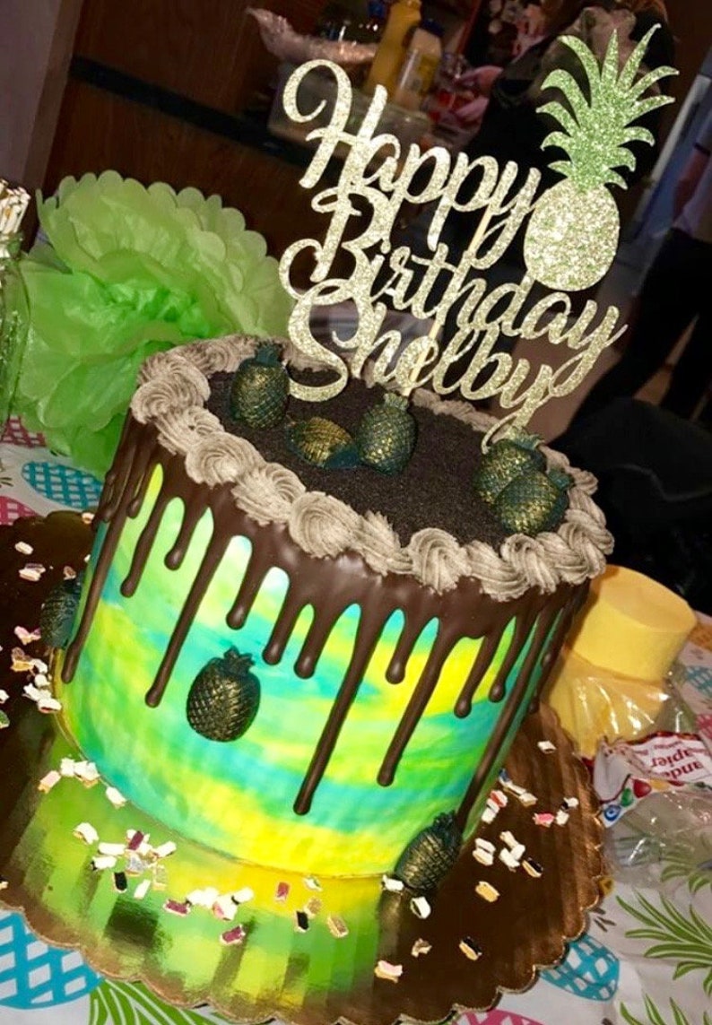 May include: A yellow and green cake with chocolate frosting and a pineapple topper. The cake is decorated with a chocolate drip and pineapple candies. The topper says "Happy Birthday Shelby".