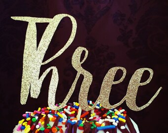 May include: Gold glitter "three" cake topper on a frosted donut with chocolate icing and colorful sprinkles. The topper is from The Paper Wish.
