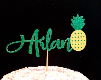 Puede incluir: Topper de pastel de purpurina verde con el nombre "Arlan" y una piña amarilla con hojas verdes.