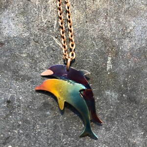 May include: A gold chain necklace with a colorful metal dolphin pendant. The dolphin is iridescent with shades of blue, green, yellow, and purple.