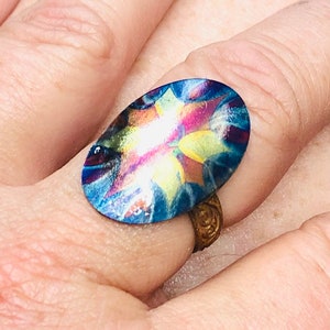 May include: A colorful oval ring with a blue, yellow, and pink design. The ring is made of metal and has a gold band.