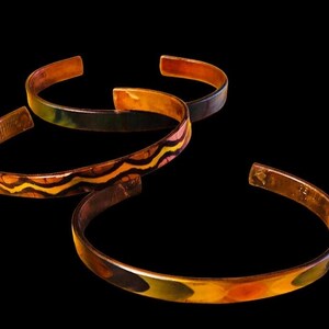 May include: Three copper bracelets with colorful enamel designs. The bracelets are stacked on top of each other, with the top bracelet having a wavy pattern in shades of orange, yellow, and black. The middle bracelet has a green and black pattern, and the bottom bracelet has a green, yellow, and orange pattern.