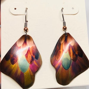 May include: A pair of handcrafted earrings with a unique, iridescent finish. The earrings feature a copper-colored metal with a feather-like design, showcasing hues of purple, green, and gold. Each earring hangs from a silver-colored hook.
