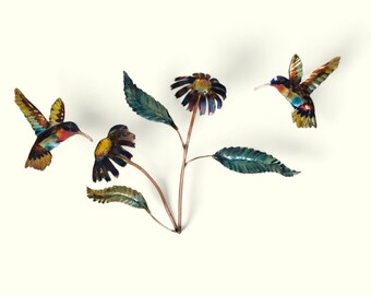May include: Metal wall art featuring two hummingbirds in flight with iridescent blue, green, and red colors. The hummingbirds are perched on a branch with three flowers with yellow, orange, and green petals.