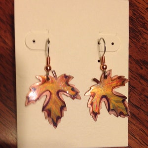 May include: A pair of leaf-shaped earrings. The earrings are copper-colored with yellow, orange, and purple accents. They hang from silver-colored hooks and are displayed on a white earring card.